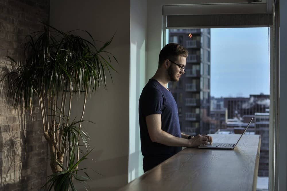 Homem em pé usando notebook no seu apartamento para trabalhar em horário flexível.